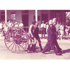 Two women walking with the Old Fire Wheel & firemen in Procession outside Courthouse Hotel, Gill Street 1967